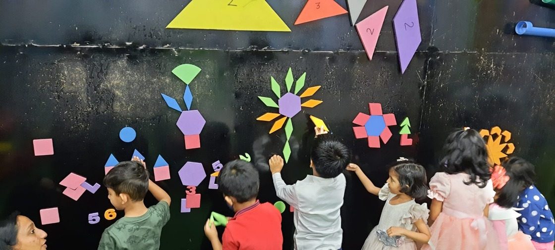 Young children building a picture out of coloured shape cut-outs on a blackboard